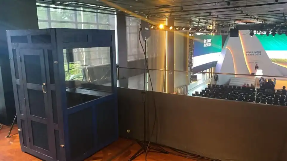 Cabine de narração azul instalada em mezanino durante evento corporativo com palco e plateia ao fundo